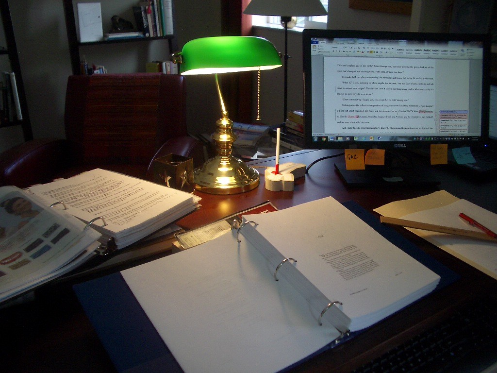 Brass desk lamp with green shade, desk, binder open to a printed manuscript